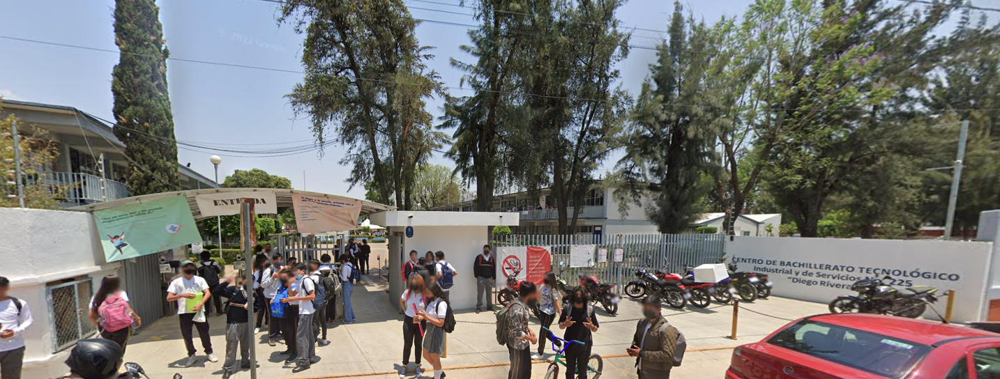 Fotografía en la que se muestra la entrada del plantel, conformada por rejas de color gris intercaladas con muros blancos. Una de las rejas, que está del lado izquierdo de la fotografía, es la que permite el ingreso a las instalaciones. Del lado derecho de la reja que da acceso hay un muro de color blanco y sobre él en letras azules dice el nombre del plantel.